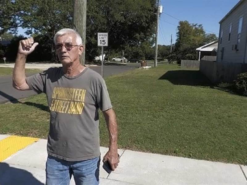 An older man with short gray hair stands on a sidewalk beside a residential street. He wears glasses, a gray T-shirt with yellow lettering that reads “SPRINGSTEEN STREET BAND,” and jeans. He gestures behind him with his thumb, as if pointing toward a location in the neighborhood under bright sunlight.