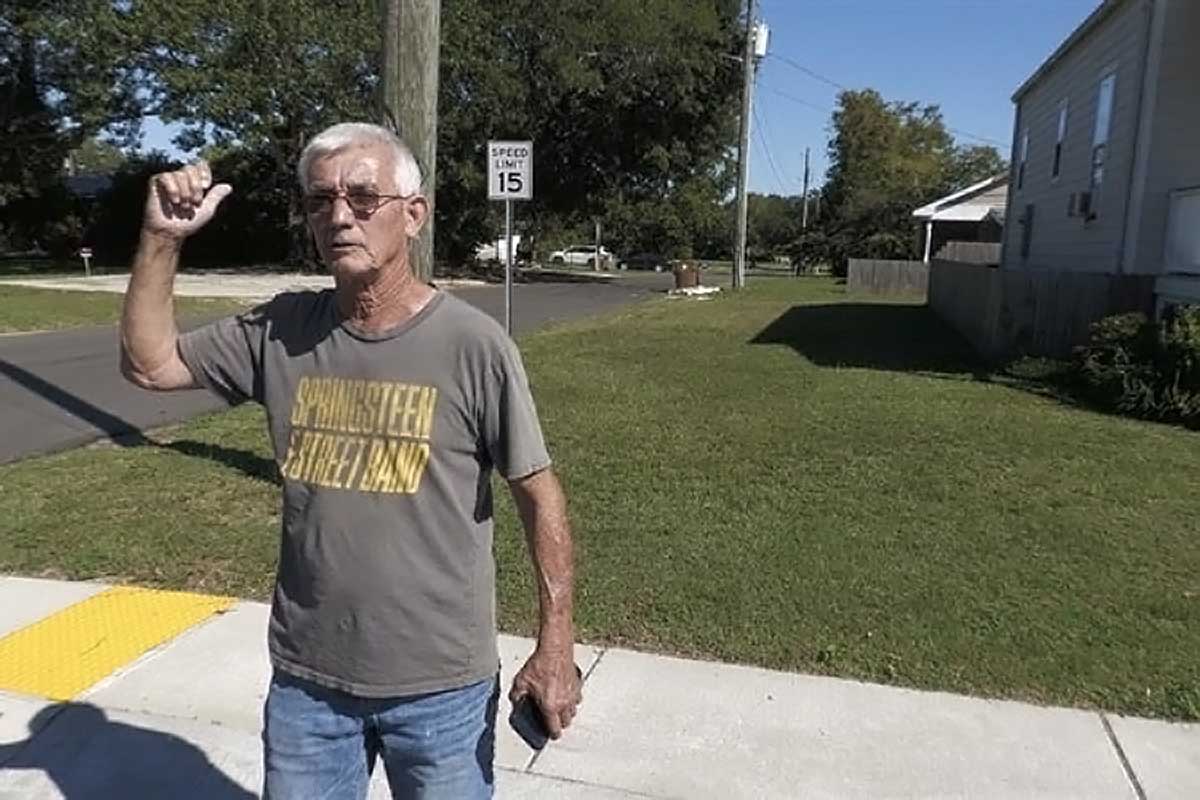 An older man with short gray hair stands on a sidewalk beside a residential street. He wears glasses, a gray T-shirt with yellow lettering that reads “SPRINGSTEEN STREET BAND,” and jeans. He gestures behind him with his thumb, as if pointing toward a location in the neighborhood under bright sunlight.