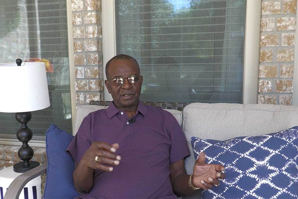 An older man wearing glasses and a purple polo shirt sits on a beige outdoor sofa on a brick porch. He gestures with his hands while speaking, framed by decorative pillows and a lamp beside him. The background shows large windows with blinds partially drawn, reflecting daylight.
