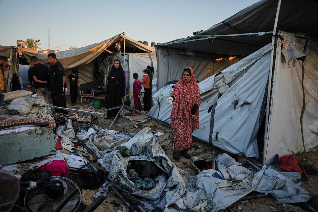 A woman in a red abaya inspects the damaged surroundings of a tent camp.