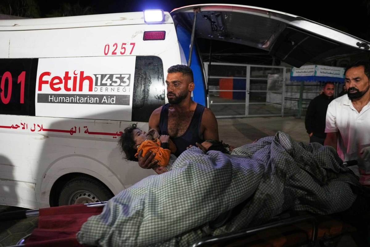 A man in a black tank top holding a young child in his arms. A gurney with a blanket is in front of him and an ambulance is behind him.