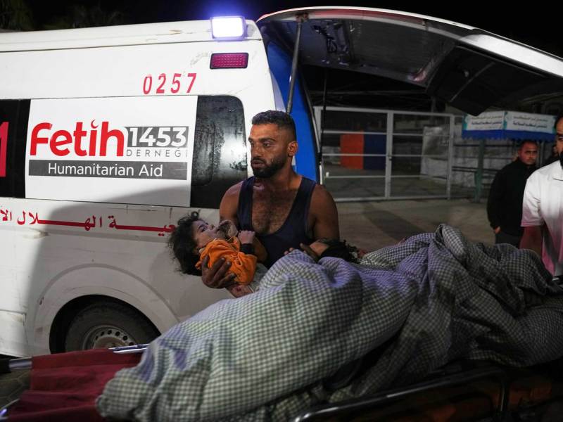 A man in a black tank top holding a young child in his arms. A gurney with a blanket is in front of him and an ambulance is behind him.