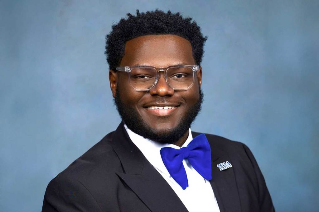 A man in a suit with blue bowtie and clear glasses smiles for a portrait against a blue background