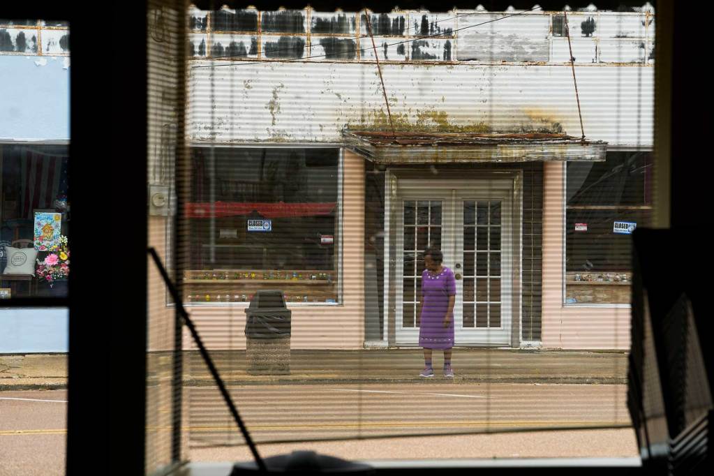 A view out a window of a woman in purple dress ready to cross the street in front of a slightly run down building