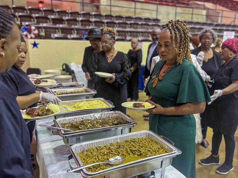 a black woman in a green dress is served beans and greens by two black women