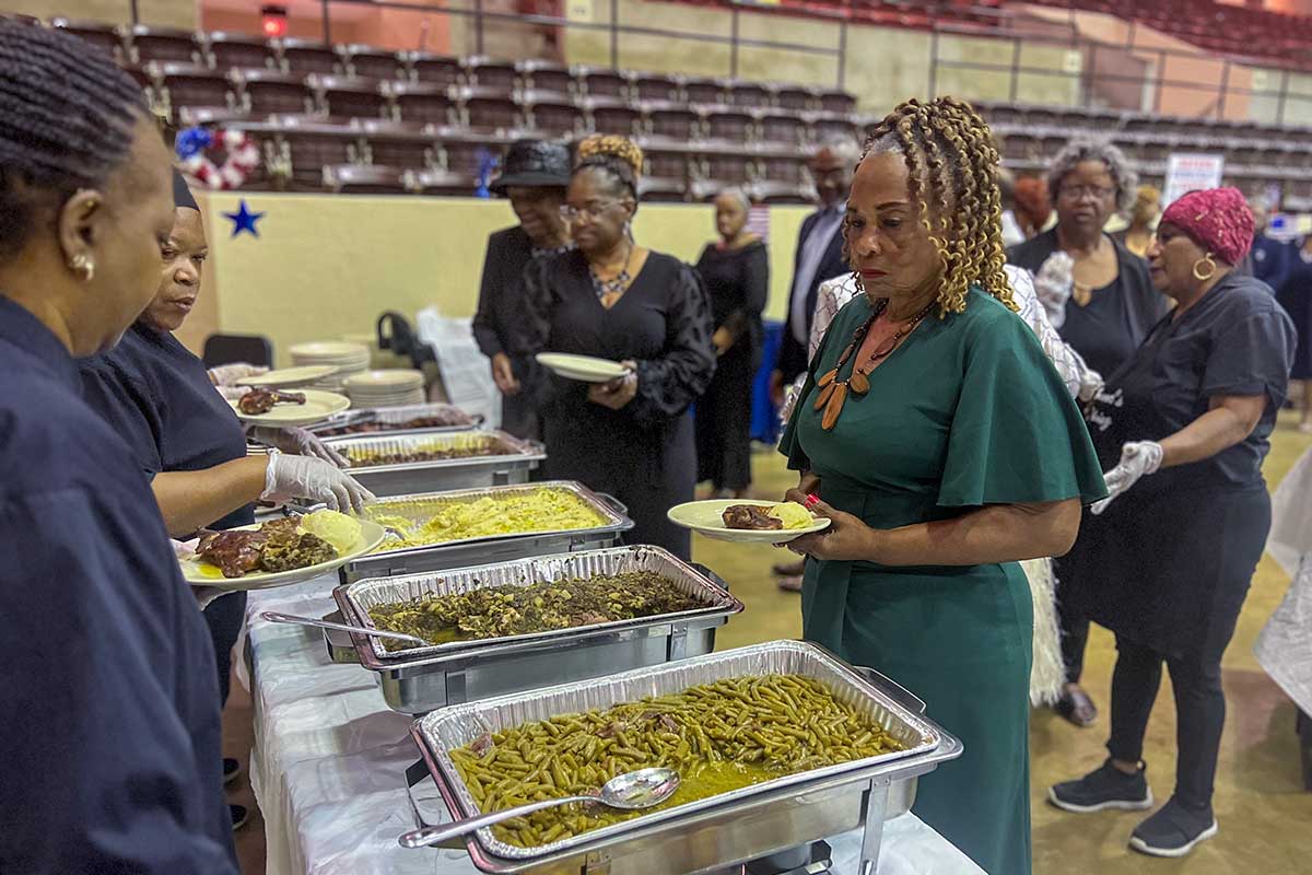 a black woman in a green dress is served beans and greens by two black women