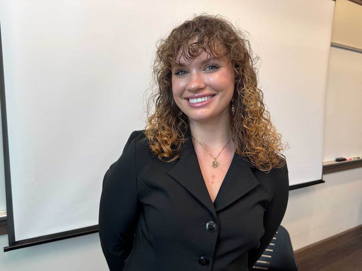 A woman with curly brown hair and wearing a black shirt smiling in front of a classroom whiteboard