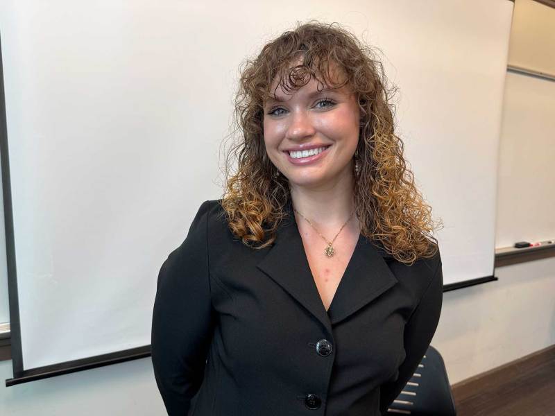 A woman with curly brown hair and wearing a black shirt smiling in front of a classroom whiteboard