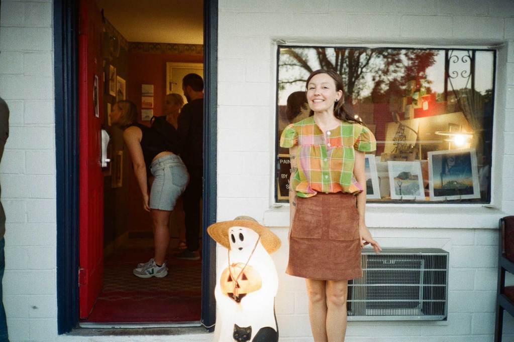 A woman stands outside of a motel room beside a halloween ghost decoration. People are visible within the doorway and art is shown in the open window.