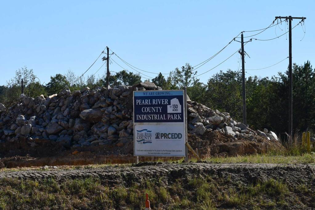 A roadside construction area with piles of gray rocks stacked behind a blue-and-white sign reading “Pearl River County Industrial Park.” Wooden utility poles and overhead wires frame the scene against a clear blue sky and a backdrop of trees.
