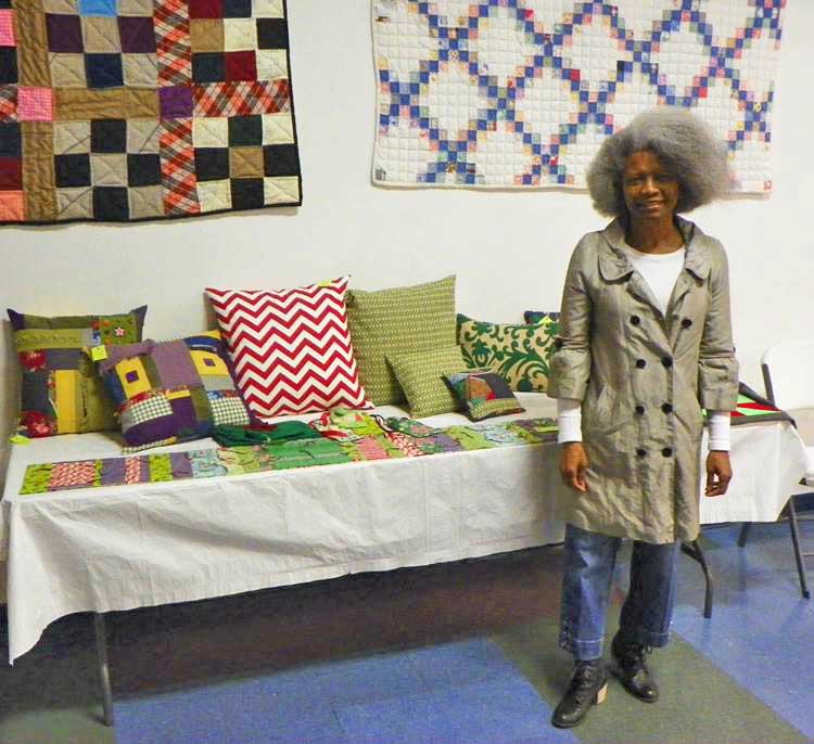 A woman stands next to a table covered in quilt and pillow art