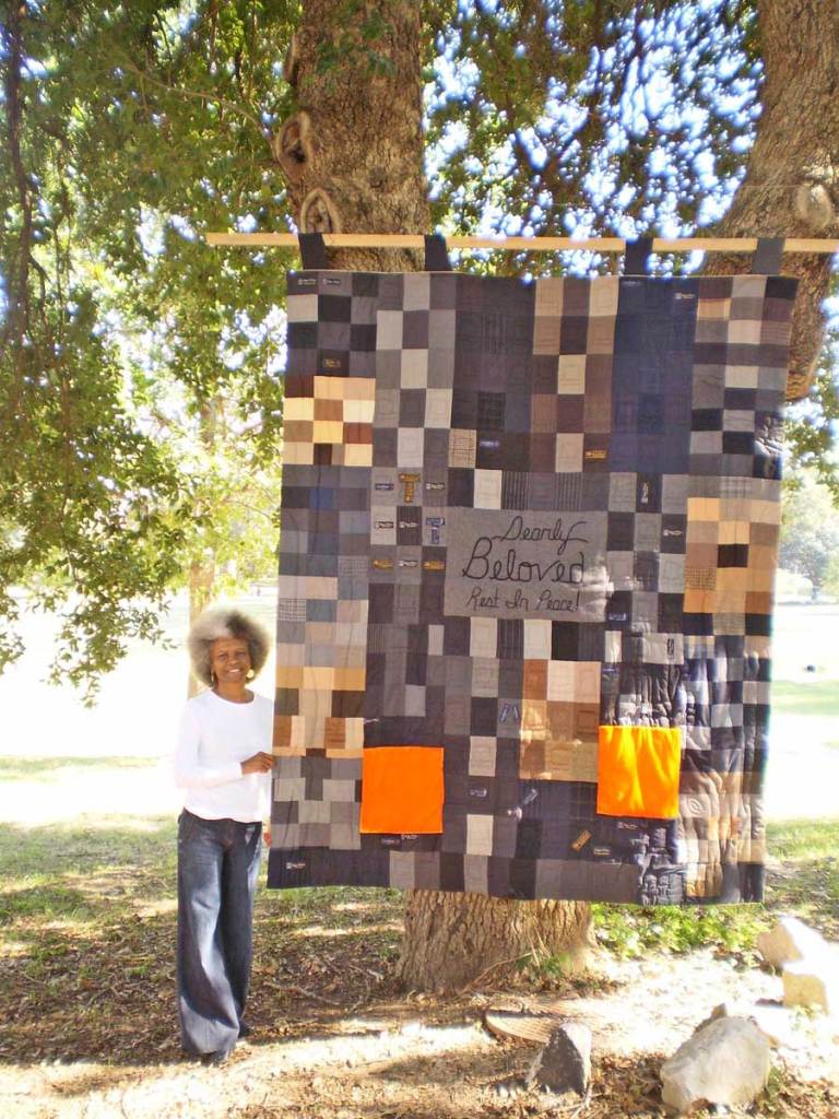 A woman stands outside next to a large dark toned quilt hanging from a tree. It reads 'Dearly Beloved Rest in Piece' in the center