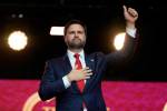 JD Vance stands on a red backdropped stage, one hand on his heart and the other making a thumbs up in the air