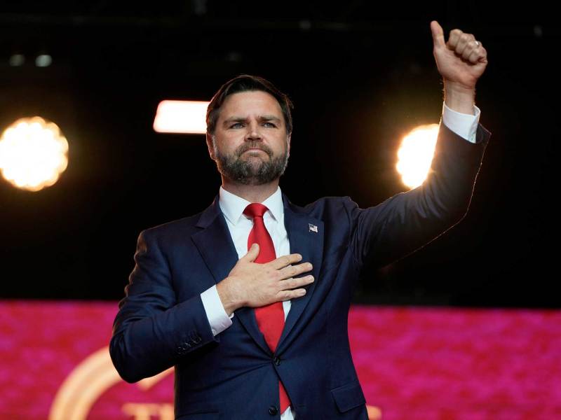JD Vance stands on a red backdropped stage, one hand on his heart and the other making a thumbs up in the air
