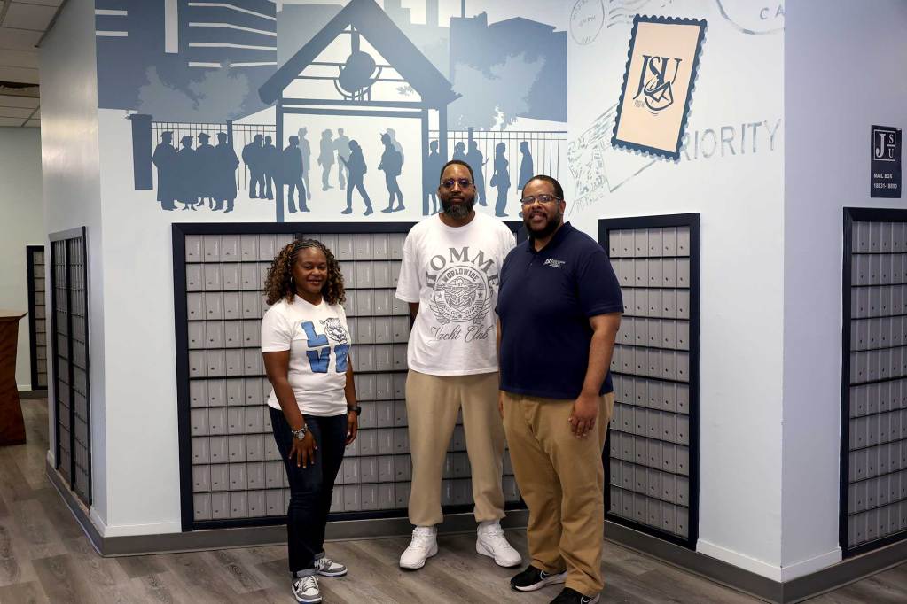 Three people pose in a mailroom , a blue mural depicting JSU students on campus seen above them