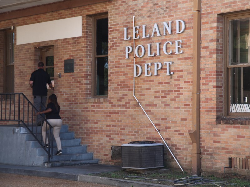 the side of a brick building is seen with two people walking up a set of steps to a door. The words "Leland Police Dept." are on the side of the building