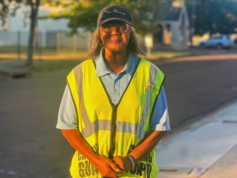 A black woman with a yellow crossing guard vest on stands with her hands clasped
