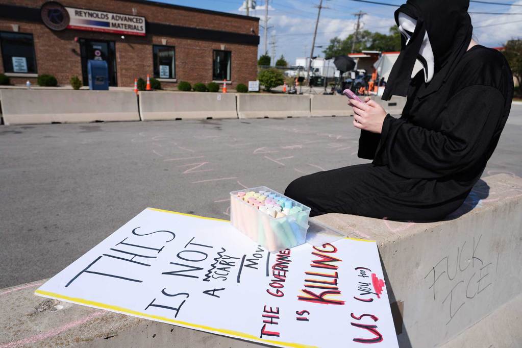 A masked protester sits on a road divider checking their phone. A sign beside them reads 'This is not a scary movie, the government is killing us (you too)'