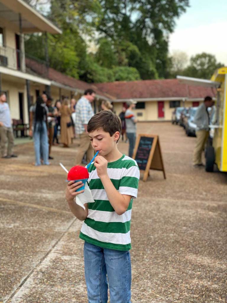 A child in a green and white stripped shirt eats a red ice cone in the parking lot of a motel being used for an art exhibit