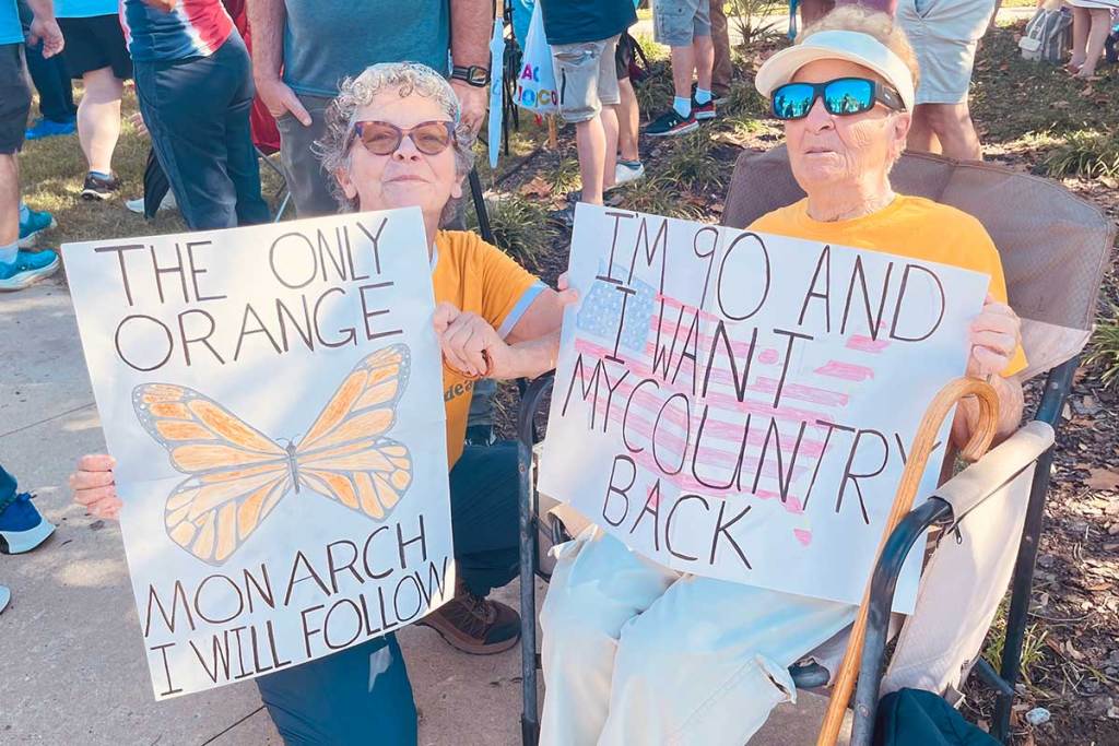Two seated protesters hold signs that read 'The only orange monarch I will follow' with a picture of a butterfly, and the other 'I am 90 and I want my country back'