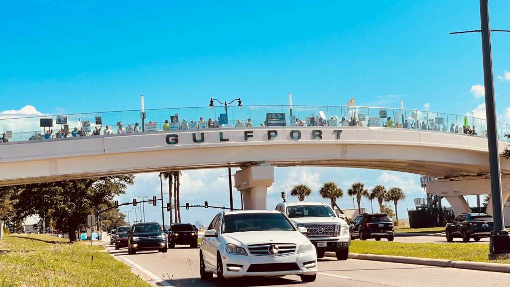 Protesters holding signs can be seen on a bridge over traffic, the bridge is labeled Gulfport