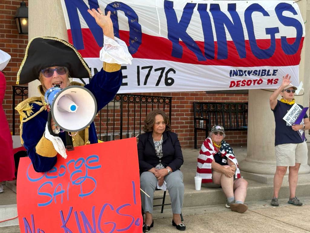 A person dressed as George Washington holds a sign that reads 'George Said No Kings!' while yelling into a megaphone