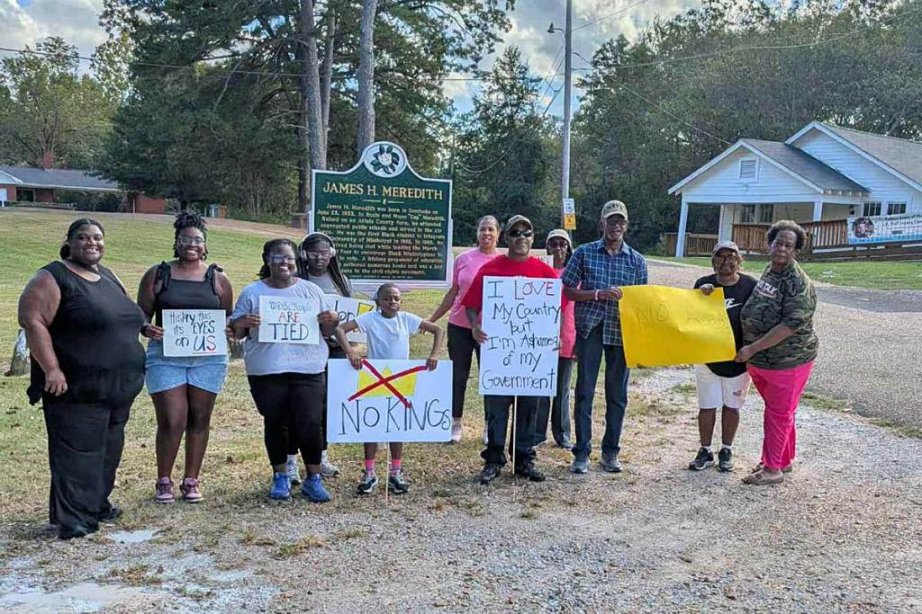 A group of protesters hold 'No Kings' signs in front of a James H. Meredith historical marker