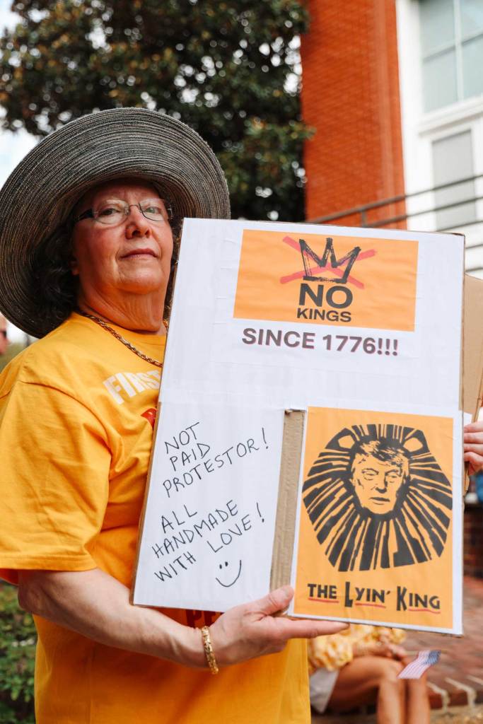 A woman holds up a sign that reads 'No kings Since 1776!!! The Lyin' King. Not paid protestor! All handmade with Love!"