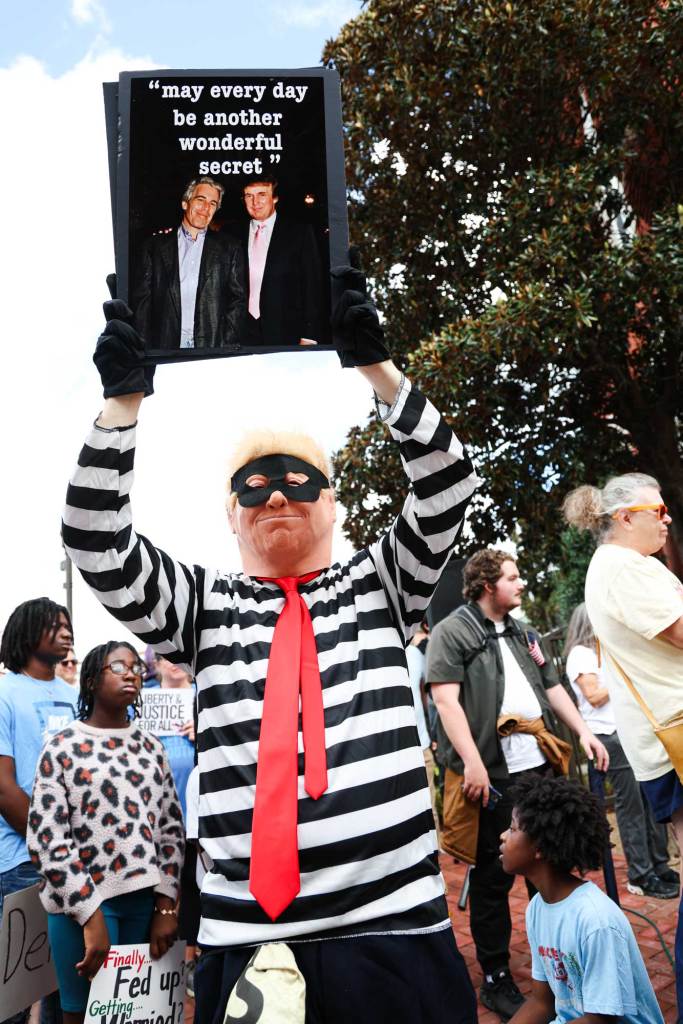A protester dressed like Donald Trump in a cartoonish burglar costume holding up a photo of Donald Trump and Jeffrey Epstein and it reads 'May every day be another wonderful secret'
