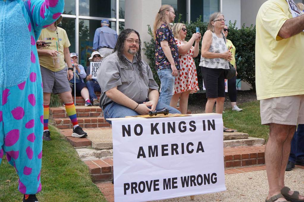 A seated man has a sign before him that reads 'No kings in America. Prove me wrong'