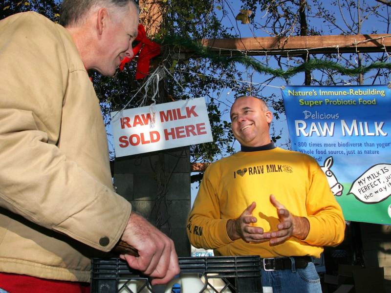 Two men stand and talk in front of signs that read Raw Milk