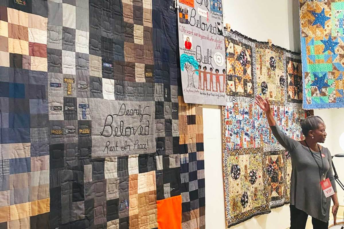 A woman gestures towards a wall filled with hanging quilts