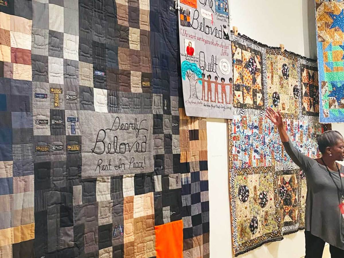 A woman gestures towards a wall filled with hanging quilts