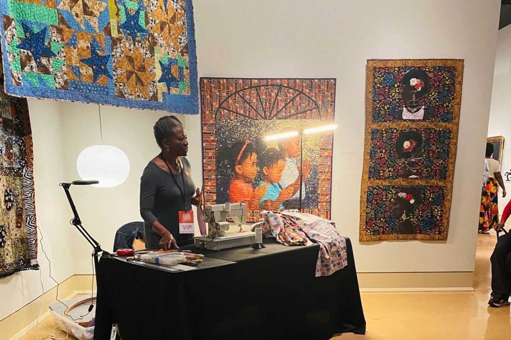 A woman stands behind a sewing table in an art museum, quilts hang on the walls behind her