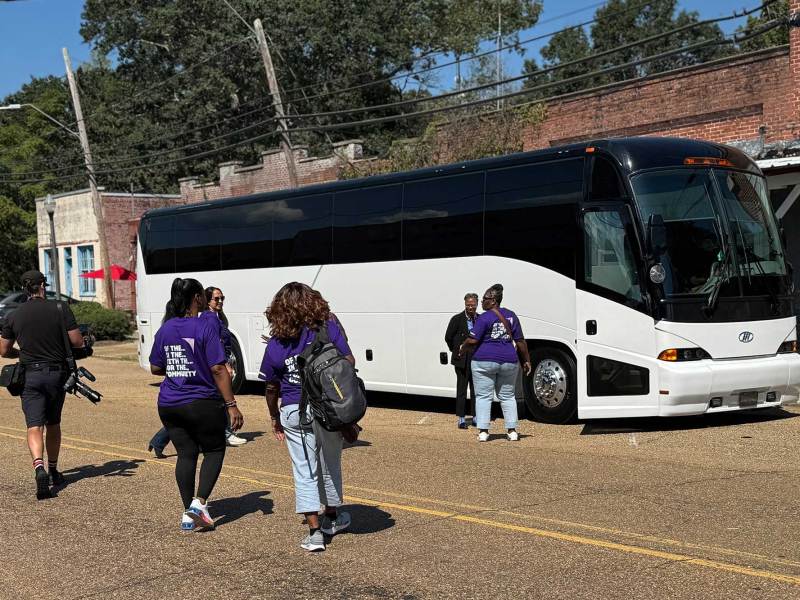 People in matching purple tshirts walk towards a black and white tour bus parked in front of a older brick building