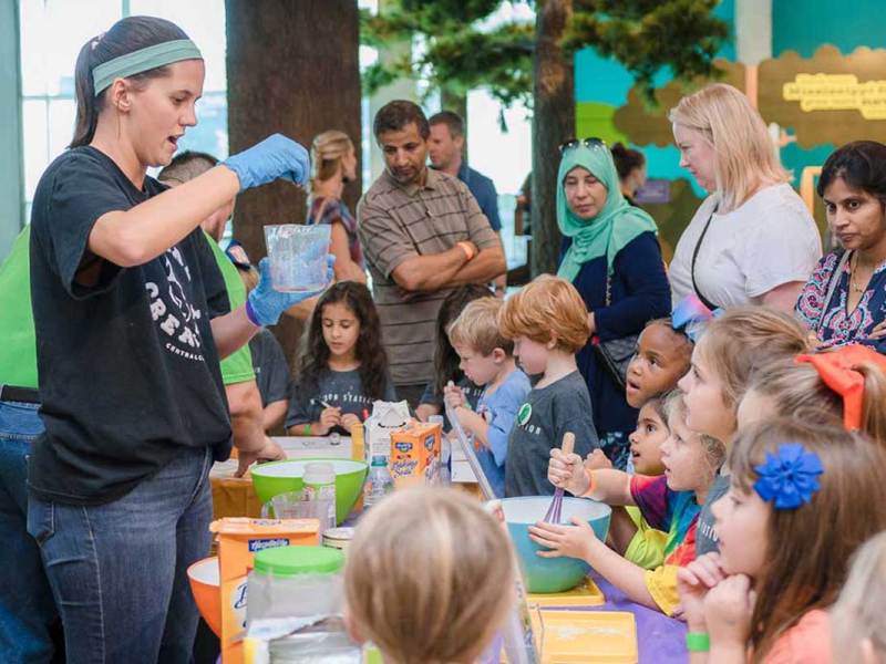 woman wearing a blue headband holds a piece of chemistry glassware while adults and children watch.