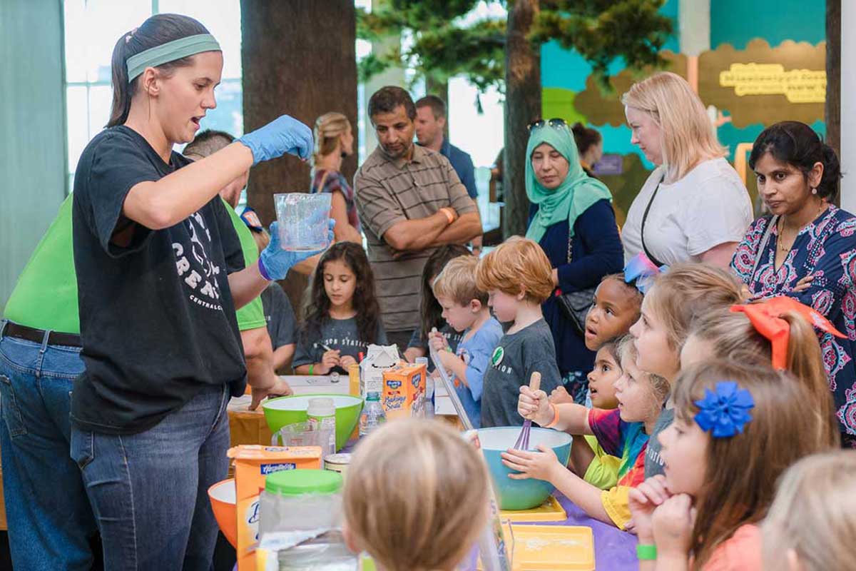 woman wearing a blue headband holds a piece of chemistry glassware while adults and children watch.