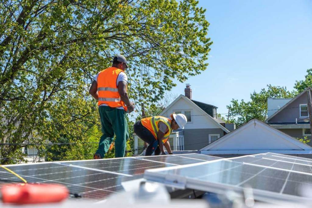 Two men in orange safety colors work on solar panels on a roof