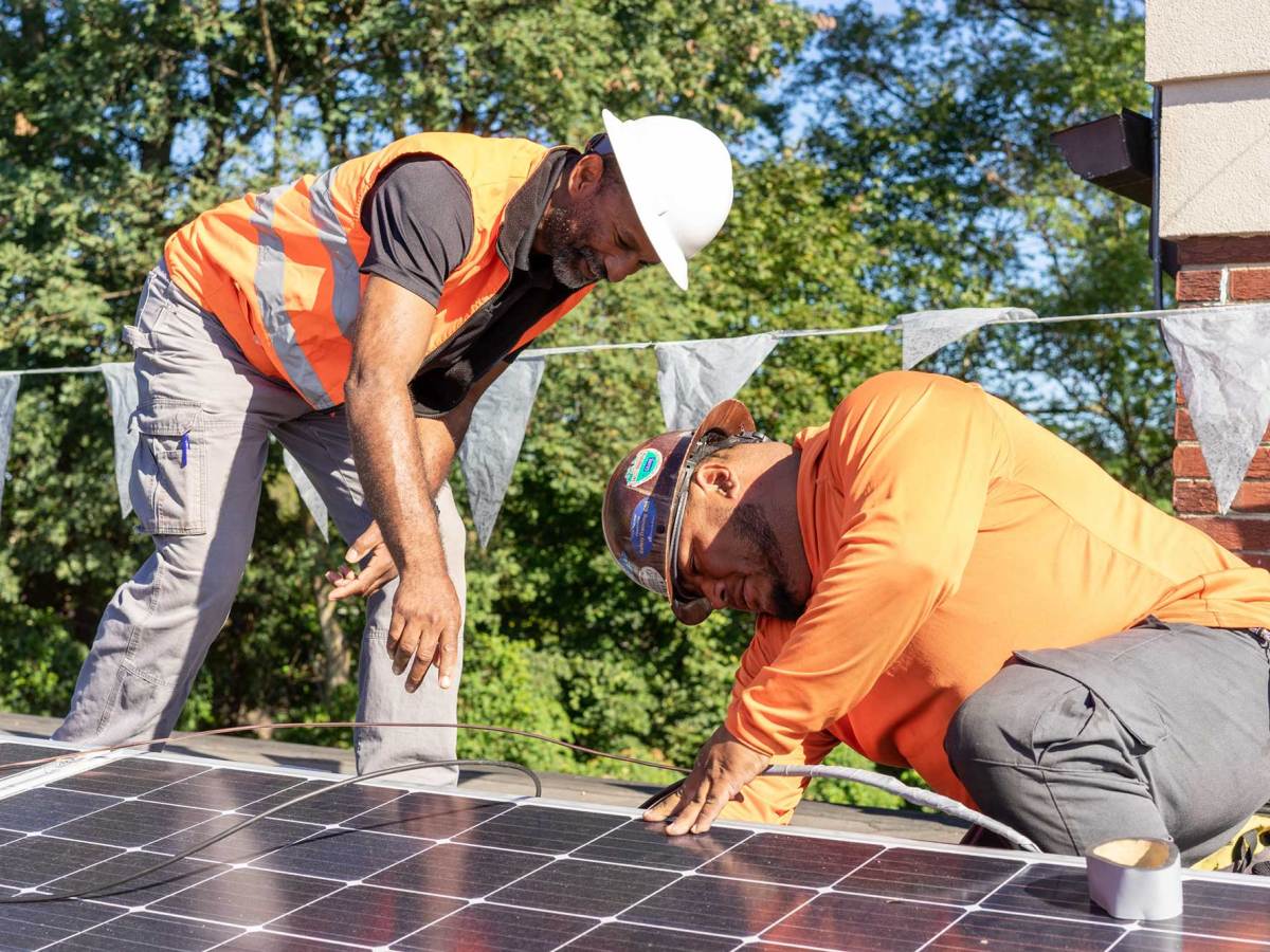 Two men in orange safety colors work on solar panels on a roof