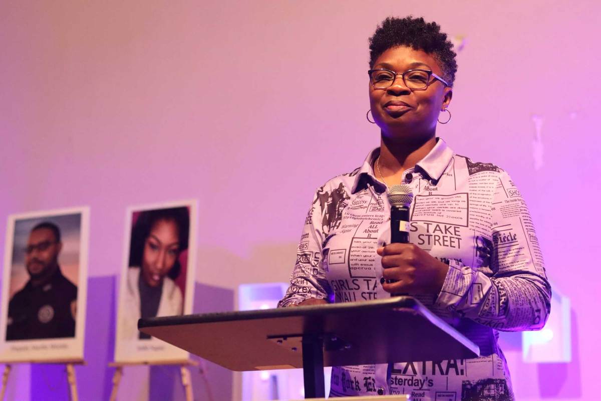 A woman holds a mic on stage, framed photos seen on easels to the left of the frame