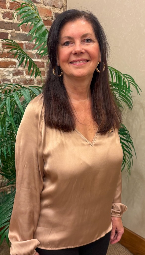 A woman in a gold blouse smiles at the camera, standing in front of a brick wall and greenery.