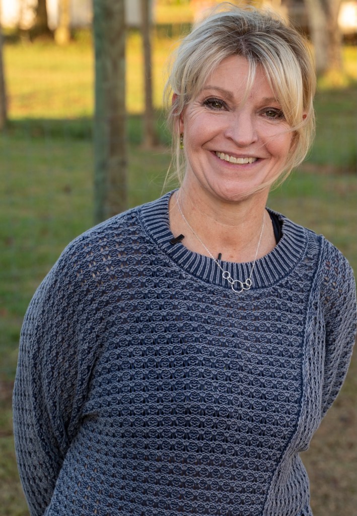 A woman wearing a blue-gray sweater stands outside, smiling.