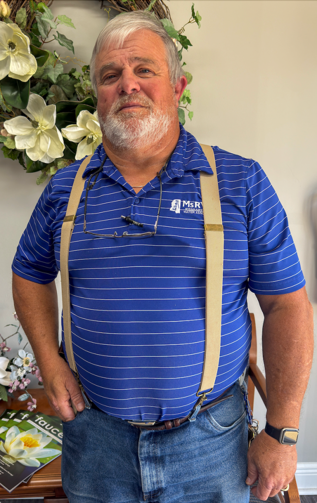A man wearing a blue striped shirt and suspenders stands indoors in front of floral decorations.