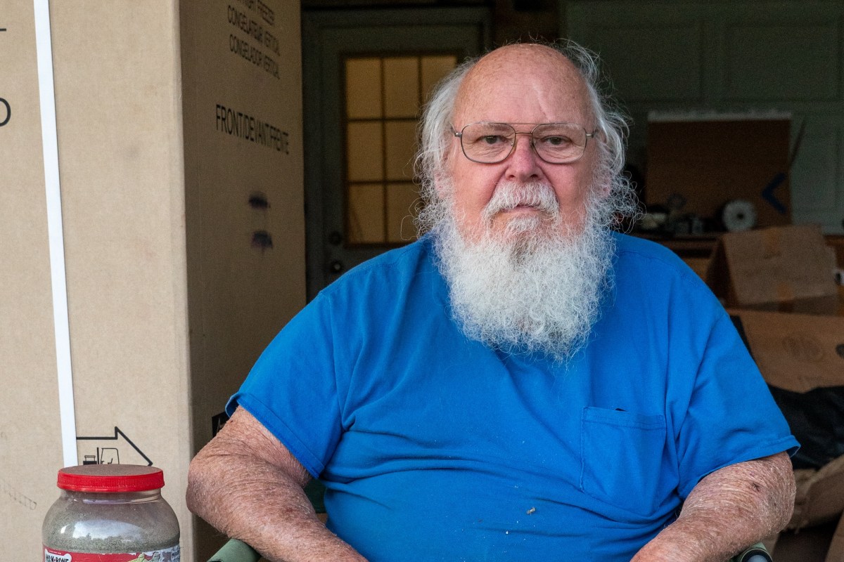 A man with a white beard wearing a blue shirt and shorts sits in a chair in his garage next to a jar of sand in a repurposed dog treat container.