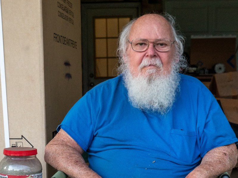 A man with a white beard wearing a blue shirt and shorts sits in a chair in his garage next to a jar of sand in a repurposed dog treat container.