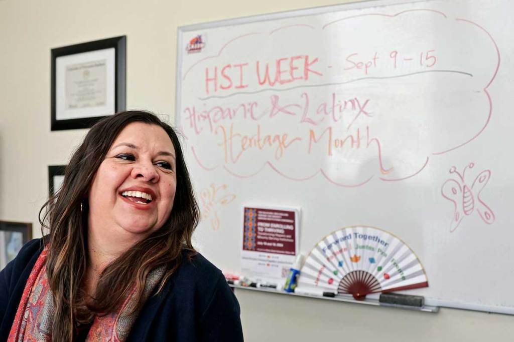 A woman with dark hair smiles and stands in front of a whiteboard that says in marker, 'HSI week, Sept 9-15