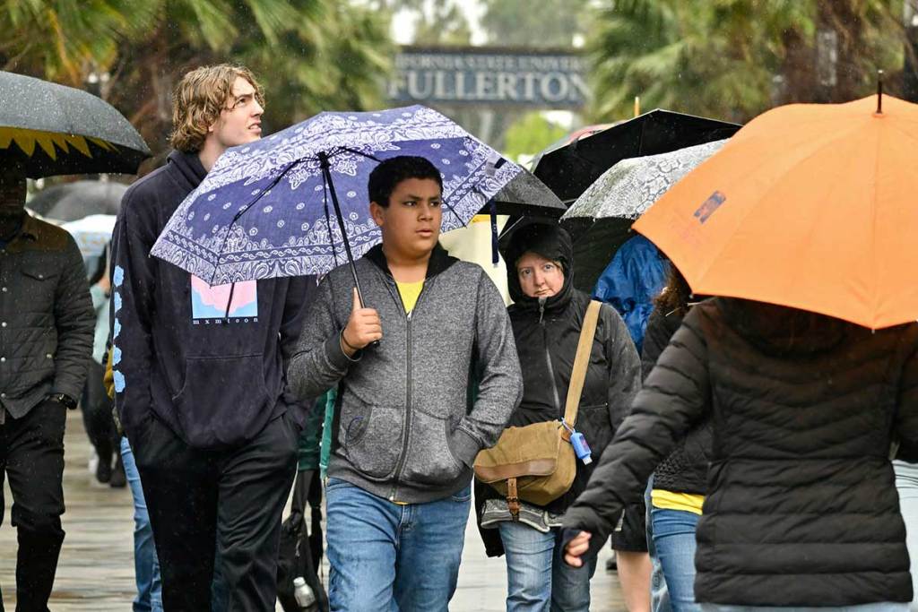 People hold umbrellas and walk together in the rain near palm trees, in front of a large sign that days Fullerton