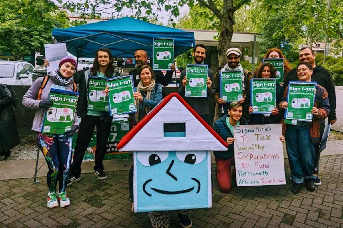 People gathered holding green signs for Seattle’s social housing initiative. Two people are dressed as house mascots.