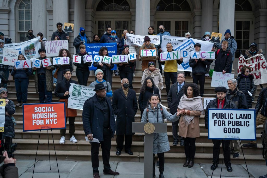 Activists stand on steps holding colorful signs while a woman stands in front of them speaking from a lectern