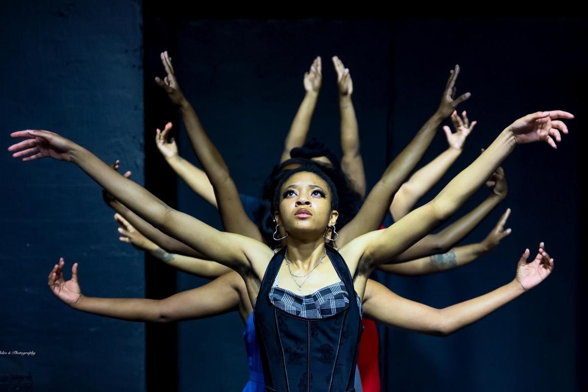 A dancer with her arms in the air, with several other dancers lined up behind her, each with their arms also extended in different positions.